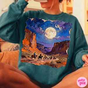 May include: Teal sweatshirt featuring a vibrant painting of a desert landscape under a full moon. The artwork depicts cowboys on horseback, riding through a canyon. The color palette includes shades of blue, orange, and purple.