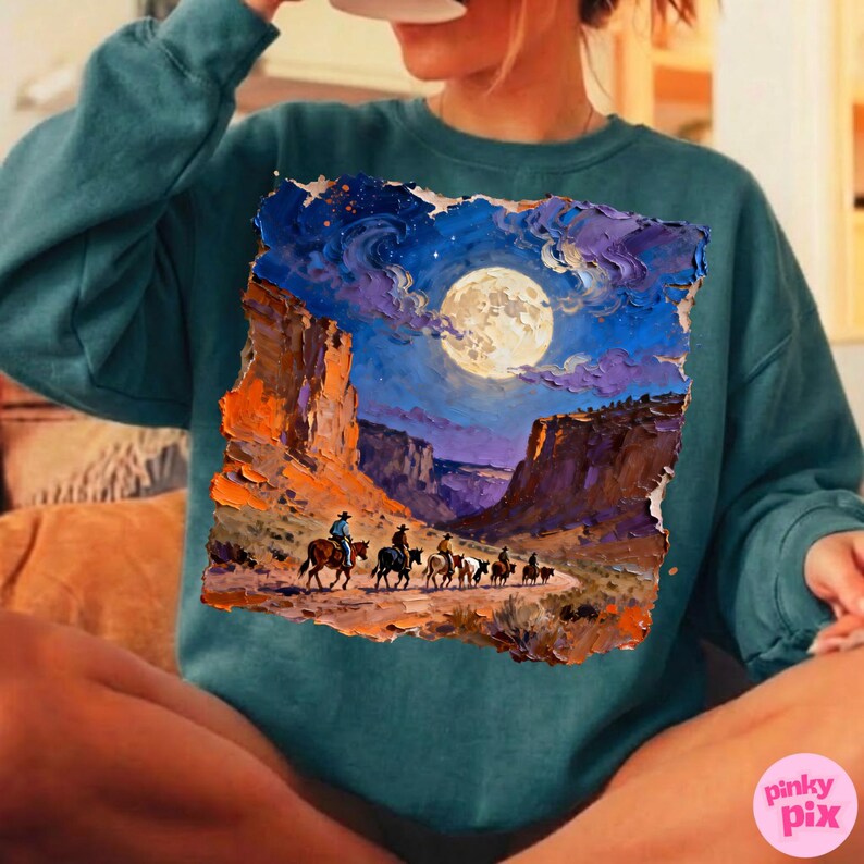 May include: Teal sweatshirt featuring a vibrant painting of a desert landscape under a full moon. The artwork depicts cowboys on horseback, riding through a canyon. The color palette includes shades of blue, orange, and purple.