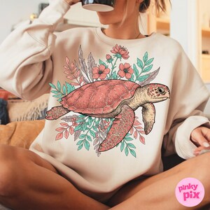 May include: A cream-colored sweatshirt with a graphic of a pink sea turtle surrounded by pink and green flowers.