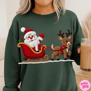May include: Dark green sweatshirt featuring a crocheted Santa Claus in a red sleigh pulled by a crocheted reindeer. Santa wears a red hat and holds a gift. The reindeer has a red harness. A festive holiday design.
