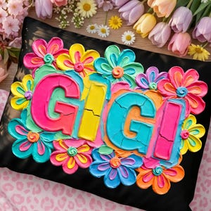 May include: Black pillow with the name "GIGI" in bright, textured letters, surrounded by colorful, raised flower designs. The flowers are in shades of pink, yellow, blue, and orange, creating a vibrant, cheerful aesthetic.