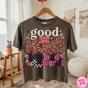 May include: A dark gray t-shirt with the phrase "good MOMS say bad words" in a playful design. The word "MOMS" features a leopard print pattern, and the design includes pink accents and floral details.