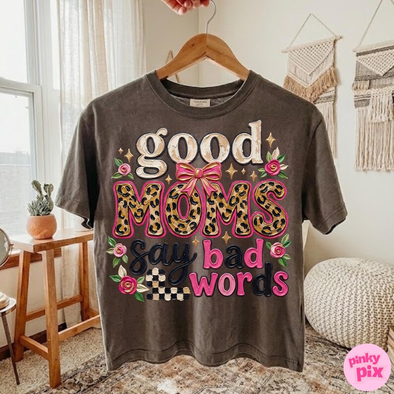 May include: A dark gray t-shirt with the phrase "good MOMS say bad words" in a playful design. The word "MOMS" features a leopard print pattern, and the design includes pink accents and floral details.