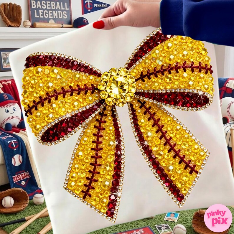 May include: A white square cushion with a large, embellished bow design. The bow is made of yellow and burgundy rhinestones, with a large central jewel. The design resembles a baseball with red stitching.