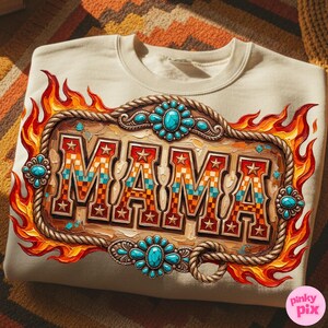 May include: Cream-colored sweatshirt featuring a decorative design with the word "MAMA" in a western-style font. The design includes a wooden-look frame, turquoise-colored accents, stars, and flames. The sweatshirt is displayed on a patterned surface.