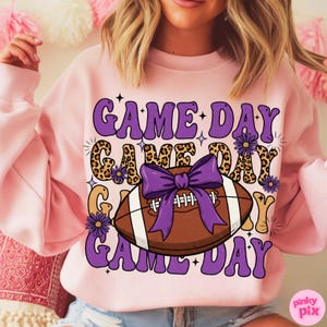 May include: Pink sweatshirt with the words "GAME DAY" in purple, with a football graphic. The football is brown with white stripes and a purple bow. The words "GAME DAY" are also in a leopard print.