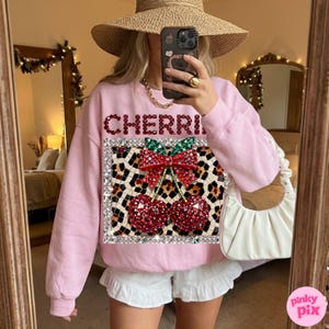 Puede incluir: Sudadera rosa claro con la palabra "CHERRI" en pedrería roja. La sudadera presenta un diseño de cereza con estampado de leopardo con cerezas de pedrería roja y un lazo verde. La imagen es de una persona que lleva la sudadera.