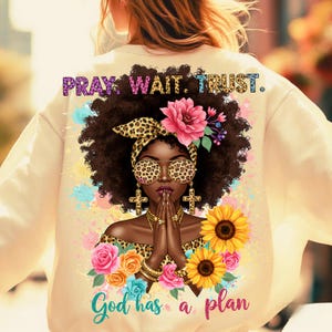 Pu&ograve; includere: Felpa color crema con grafica di una donna nera con fascia e occhiali da sole leopardati, mani giunte in preghiera e dettagli floreali. Il testo "PRAY. WAIT. TRUST." e "God has a plan" sono visibili.