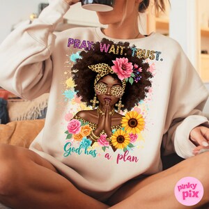 May include: A beige sweatshirt with a graphic print of a Black woman with a leopard print headband, praying with her hands clasped. The graphic also includes flowers, sunflowers, and the text "Pray. Wait. Trust. God has a plan."
