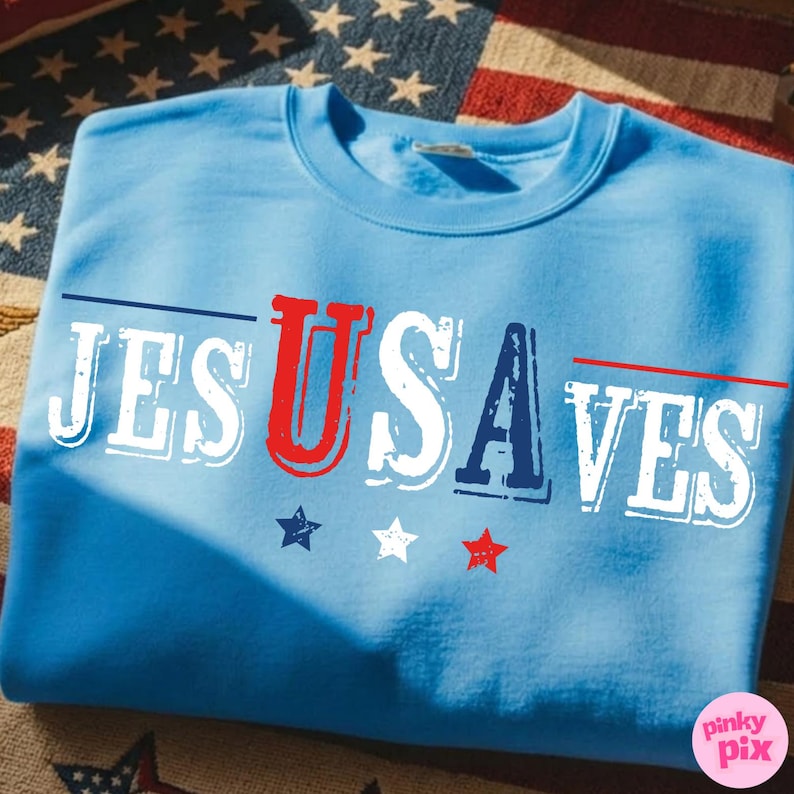 May include: A light blue sweatshirt with the words "JESUS USA SAVES" printed across the front in red, white, and blue. The design includes star graphics. The sweatshirt is folded on a surface with an American flag pattern.
