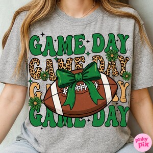 May include: Gray t-shirt with a football graphic and the words "GAME DAY" in green, with a leopard print design. The football has a green bow. The shirt is a casual, sporty style.