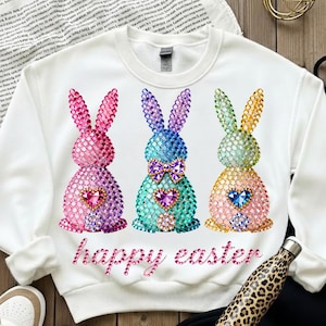 May include: White sweatshirt featuring three jeweled bunny designs in pink, blue, and yellow, each with a heart-shaped gem. The words "happy easter" are written in pink script below the bunnies. A leopard print water bottle is in the bottom right corner.