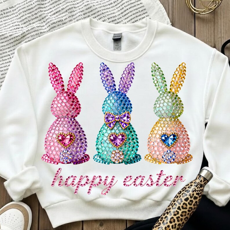 May include: White sweatshirt featuring three jeweled bunny designs in pink, blue, and yellow, each with a heart-shaped gem. The words "happy easter" are written in pink script below the bunnies. A leopard print water bottle is in the bottom right corner.