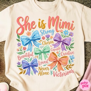 K&ouml;nnte beinhalten: Cremefarbenes T-Shirt mit dem Text "She is Mimi" in rosa und gelber Schrift. Das Design umfasst bunte Schleifen, Blumen und W&ouml;rter wie "Strong", "Chosen" und "Victorious".
