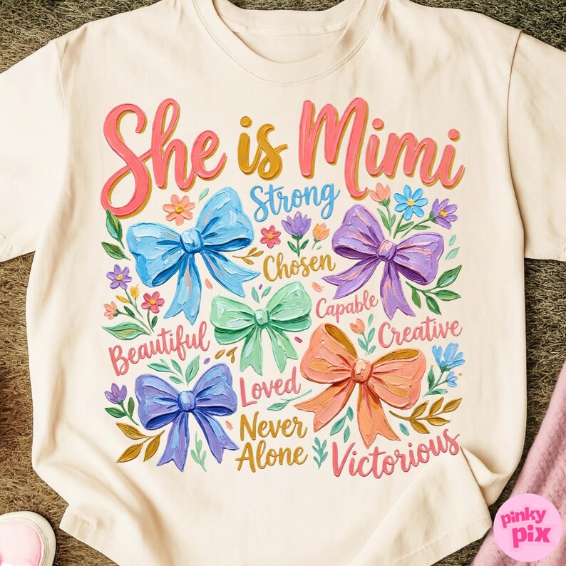 K&ouml;nnte beinhalten: Cremefarbenes T-Shirt mit dem Text "She is Mimi" in rosa und gelber Schrift. Das Design umfasst bunte Schleifen, Blumen und W&ouml;rter wie "Strong", "Chosen" und "Victorious".