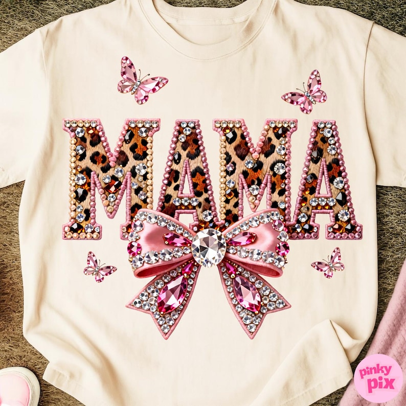May include: Cream-colored t-shirt featuring the word "MAMA" in a leopard print design, outlined with pink rhinestones. A pink bow with a large crystal center and pink gems is below the word. Pink butterflies are scattered around the design.
