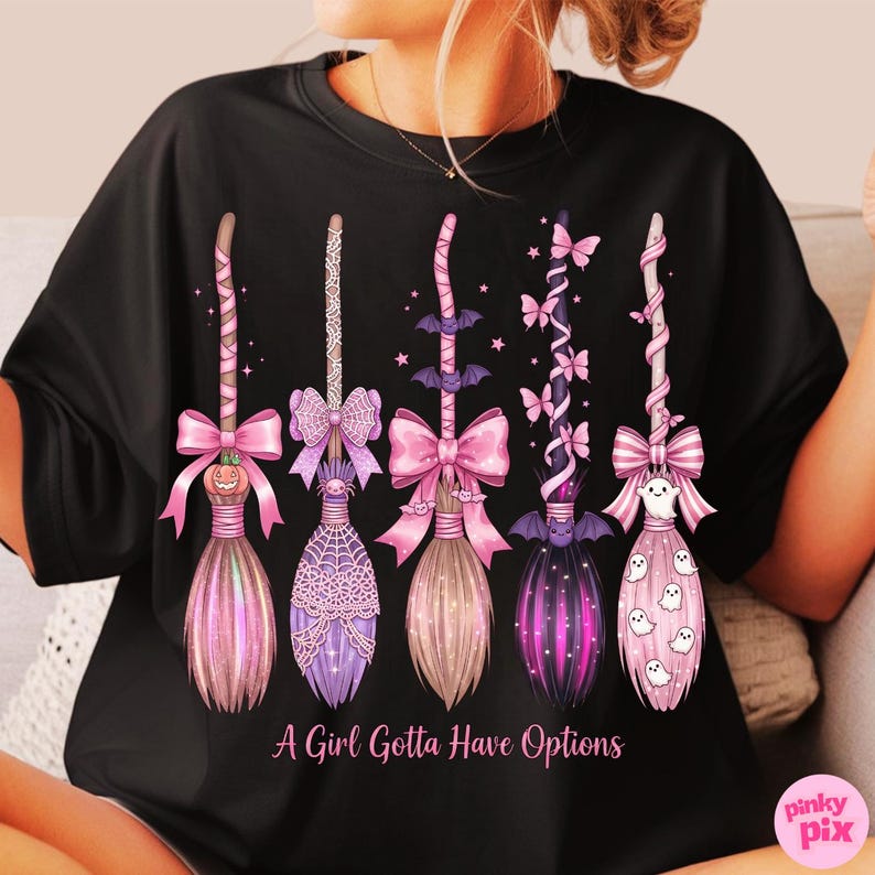 May include: Black t-shirt featuring five decorative witch brooms in shades of pink, purple, and black. Each broom is adorned with bows, bats, and ghosts. The text "A Girl Gotta Have Options" is printed below the brooms.