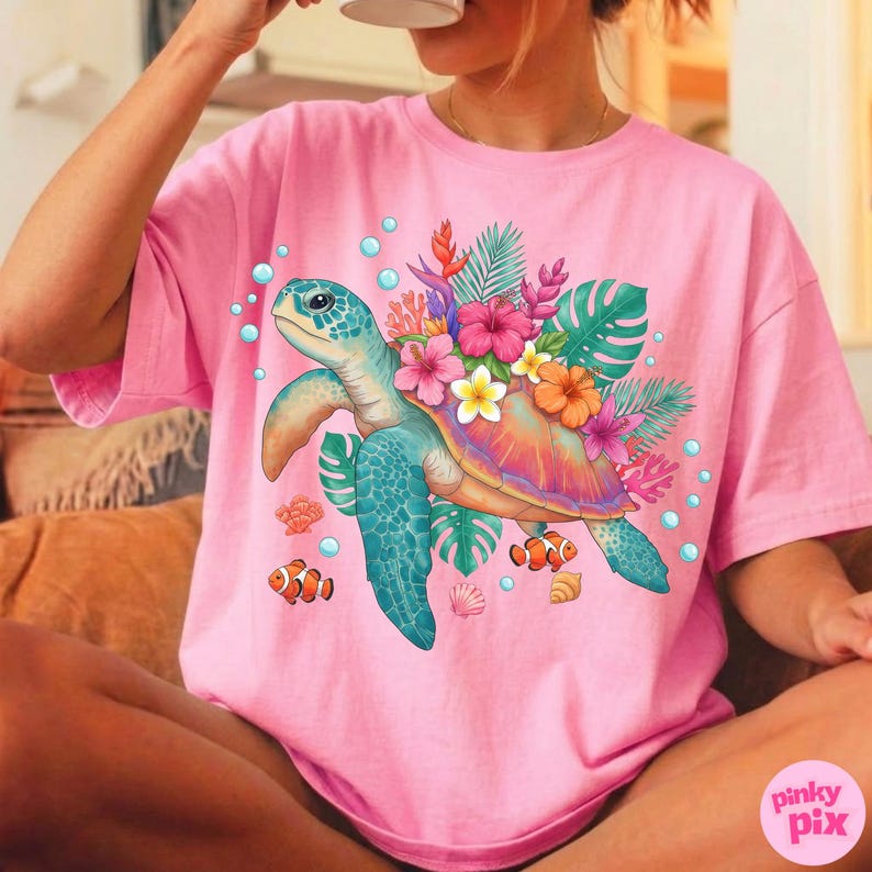 May include: Pink t-shirt featuring a colorful sea turtle design. The turtle has a blue shell and is adorned with tropical flowers and leaves. Bubbles and small fish surround the turtle, creating a vibrant, summery look.