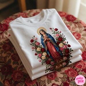 May include: A stack of white t-shirts featuring a religious illustration. The design depicts a woman in a blue and red robe with a gold halo, surrounded by red, pink, and yellow roses. The shirts are neatly folded.