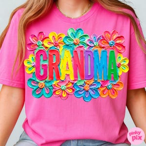 May include: A pink t-shirt featuring the word "GRANDMA" in colorful, 3D-style letters, surrounded by vibrant, multi-colored flower designs. The shirt has short sleeves and a crew neck.