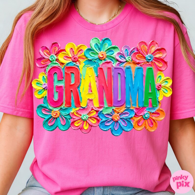 May include: A pink t-shirt featuring the word "GRANDMA" in colorful, 3D-style letters, surrounded by vibrant, multi-colored flower designs. The shirt has short sleeves and a crew neck.