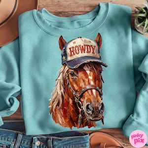 May include: A light blue sweatshirt featuring a detailed illustration of a brown horse wearing a "HOWDY" cap. The horse has a tan mane and is wearing a bridle. The cap is navy blue with gold stars and the word "HOWDY" in red.