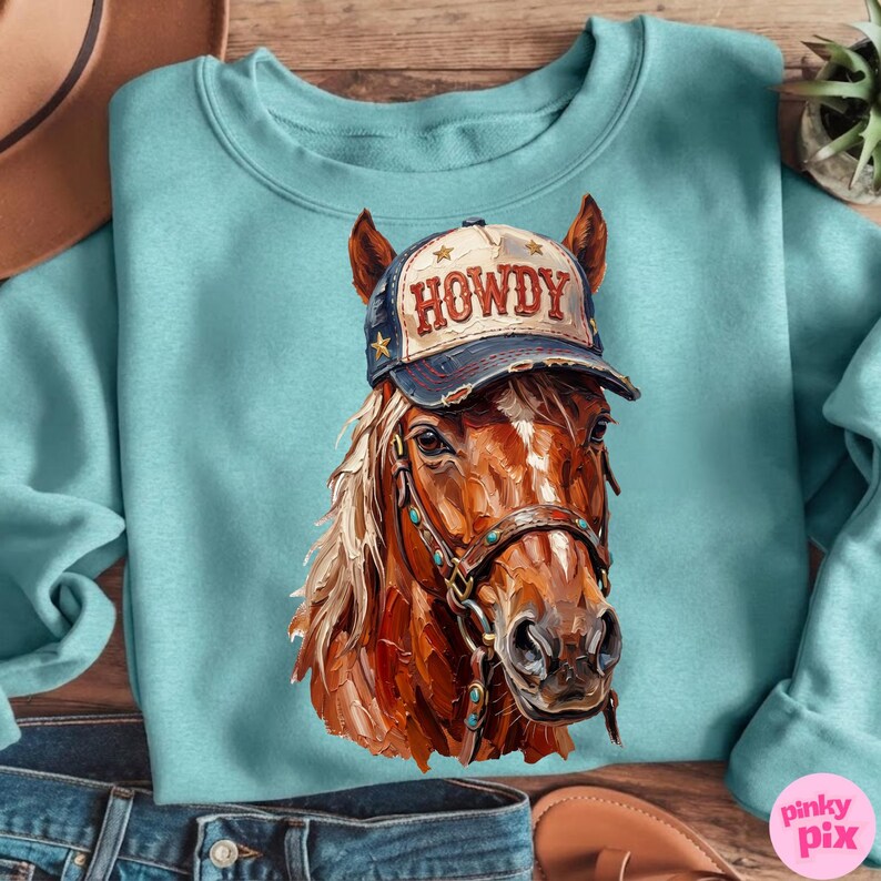 May include: A light blue sweatshirt featuring a detailed illustration of a brown horse wearing a "HOWDY" cap. The horse has a tan mane and is wearing a bridle. The cap is navy blue with gold stars and the word "HOWDY" in red.