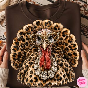 May include: A dark brown sweatshirt featuring a graphic print of a turkey with leopard print feathers, wearing gold-rimmed glasses and a large, multi-faceted clear crystal statement necklace. The turkey has a red wattle and a sharp beak.