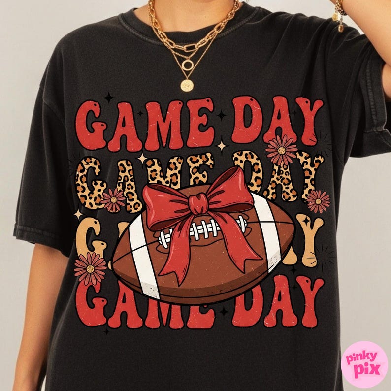May include: Black t-shirt with a retro design featuring the words "GAME DAY" in red, with a leopard print accent. A football with a red bow is in the center. The shirt has a vintage, worn look.