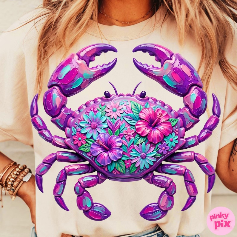 May include: A light beige t-shirt featuring a vibrant, stylized crab design. The crab is rendered in shades of purple, pink, and teal, with intricate floral details filling its body. The design includes the text "pinky pix" in the lower right corner.
