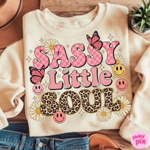 May include: Cream-colored sweatshirt with the text "SASSY Little SOUL" in pink, checkered, and leopard print lettering. Features butterflies, daisies, and smiley faces. A fun, retro-style graphic.