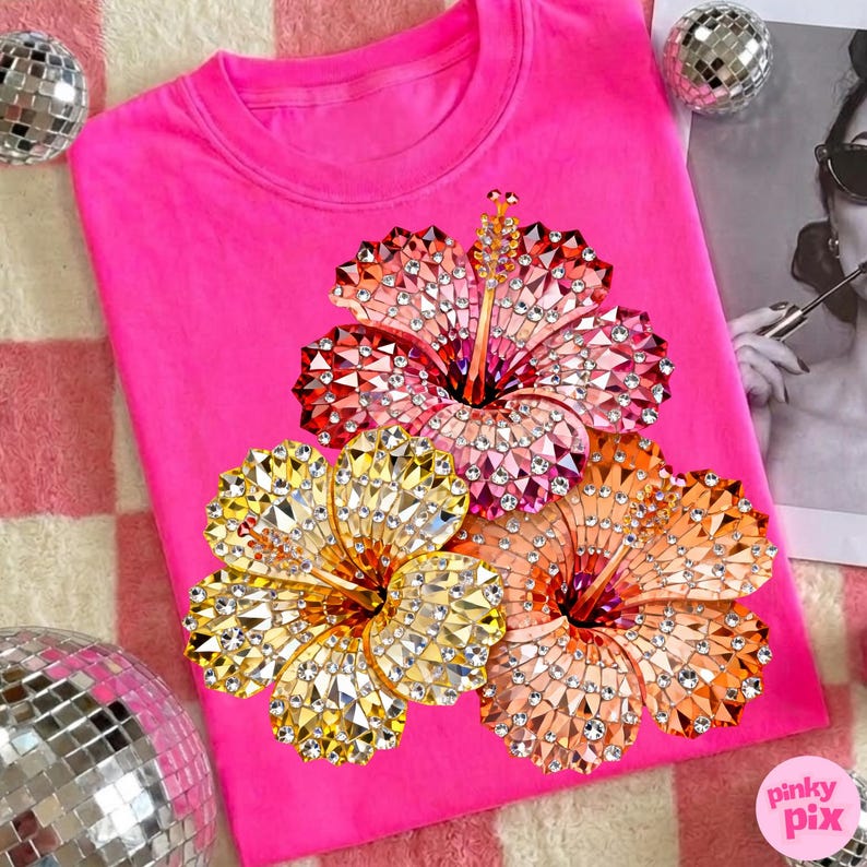 May include: A bright pink sweatshirt featuring three jeweled hibiscus flowers in shades of red, gold, and orange. The flowers are embellished with sparkling rhinestones. The sweatshirt is on a pink and white checkered surface.