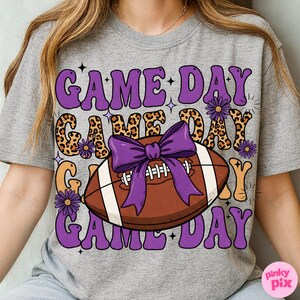 May include: Gray t-shirt with a football graphic and the words "GAME DAY" in purple, leopard print, and orange. A purple bow is on the football. The shirt has a retro, groovy style.