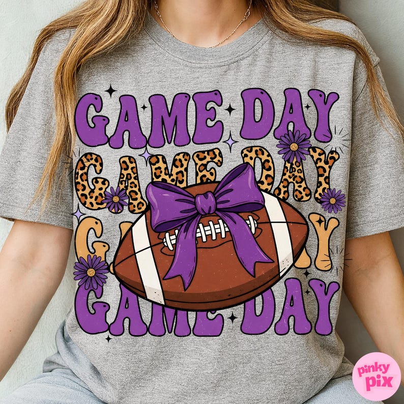 May include: Gray t-shirt with a football graphic and the words "GAME DAY" in purple, leopard print, and orange. A purple bow is on the football. The shirt has a retro, groovy style.