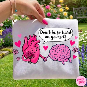 May include: White square pouch with a pink cartoon heart holding flowers and a pink cartoon brain holding hands. The text "Don't be so hard on yourself" is in a speech bubble. Pink hearts and flowers decorate the pouch.