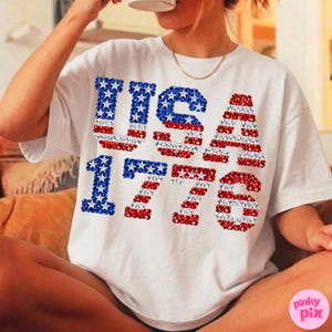K&ouml;nnte beinhalten: Wei&szlig;es T-Shirt mit dem Text "USA 1776" in einem patriotischen Design. Die Buchstaben und Zahlen sind mit roten, wei&szlig;en und blauen Mustern gef&uuml;llt, die an die amerikanische Flagge erinnern. Ein l&auml;ssiges T-Shirt.