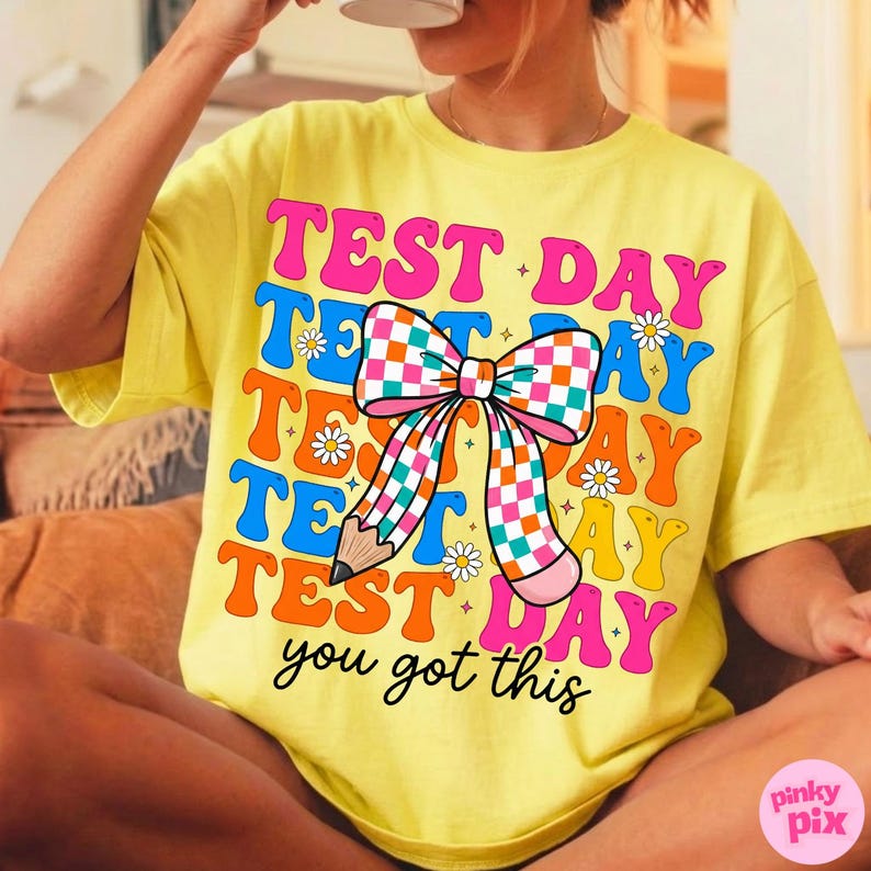 May include: Yellow t-shirt with the words "TEST DAY" in a retro font, with a large bow and pencil graphic. The shirt also includes the phrase "you got this". The design features pink, blue, orange, and yellow colors.