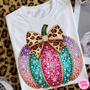 Op de afbeelding: Een wit t-shirt met een grote pompoenafbeelding versierd met kleurrijke strass-steentjes in roze, turquoise en paarse tinten. De pompoen is voorzien van een strik met luipaardprint en een groene steel, alles bezet met fonkelende kristallen.