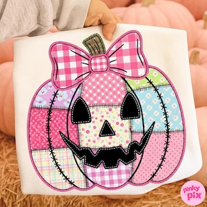 May include: A white tote bag featuring a patchwork pumpkin design. The pumpkin has a black jack-o'-lantern face and is decorated with pink, blue, and yellow patterned fabric. A pink gingham bow sits atop the pumpkin.