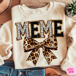 May include: Cream-colored sweatshirt with the word "MEME" in a jeweled font, outlined in gold and black. A large leopard print bow with gold accents is centered below the text. The sweatshirt is a casual, stylish garment.