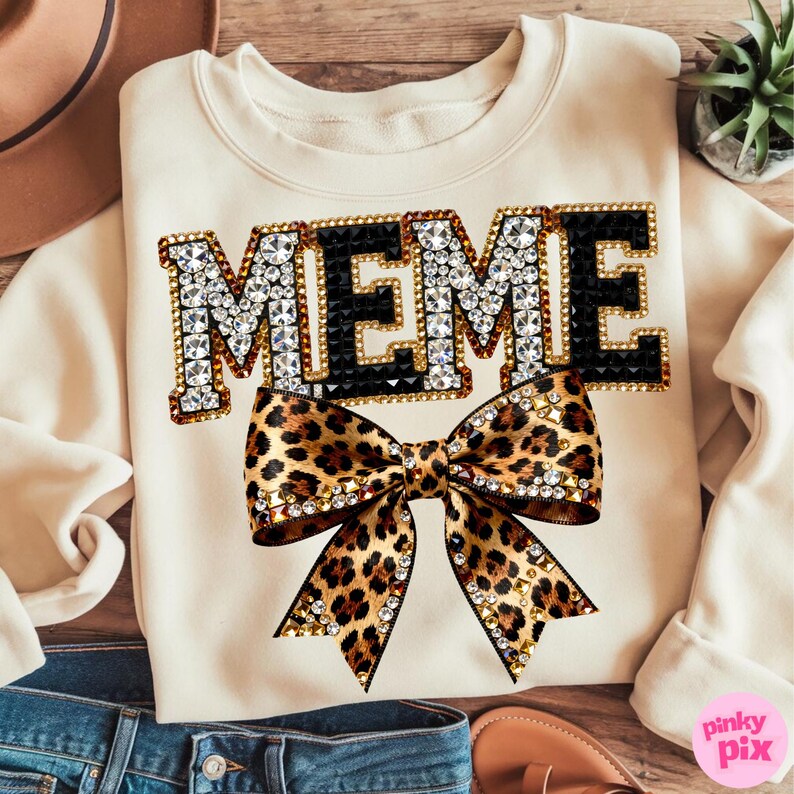 May include: Cream-colored sweatshirt with the word "MEME" in a jeweled font, outlined in gold and black. A large leopard print bow with gold accents is centered below the text. The sweatshirt is a casual, stylish garment.