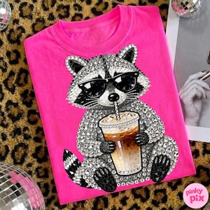 May include: A hot pink t-shirt featuring a rhinestone-studded raccoon wearing sunglasses and holding an iced coffee. The raccoon design is centered on the shirt. The shirt has a crew neck.