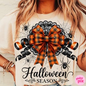 May include: Beige t-shirt with a Halloween theme. The shirt features a large orange and black plaid bow, black lace, spiderwebs, and spiders. The words "Halloween Season" are written in black script.