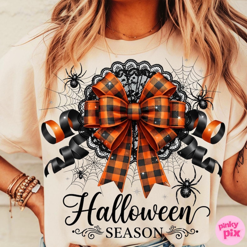 May include: Beige t-shirt with a Halloween theme. The shirt features a large orange and black plaid bow, black lace, spiderwebs, and spiders. The words "Halloween Season" are written in black script.