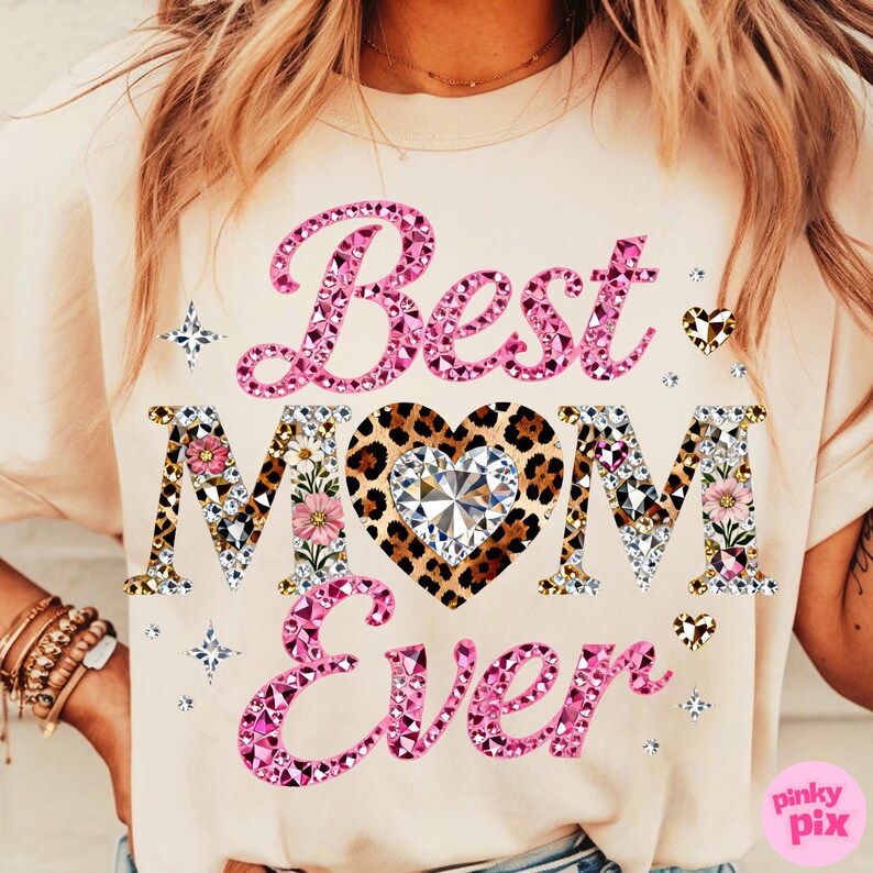 May include: A light beige t-shirt with the text "Best Mom Ever" in pink, leopard print, and crystal embellishments. The word "Mom" is in a leopard print heart shape with a crystal center. Floral and star accents complete the design.
