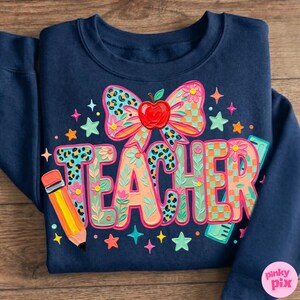 May include: Navy blue sweatshirt with a colorful "TEACHER" graphic. The design includes a bow with an apple, a pencil, stars, and floral accents. The letters have leopard print and checkered patterns.