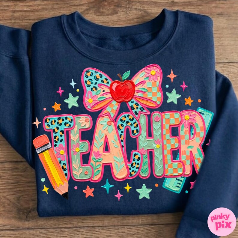 May include: Navy blue sweatshirt with a colorful "TEACHER" graphic. The design includes a bow with an apple, a pencil, stars, and floral accents. The letters have leopard print and checkered patterns.