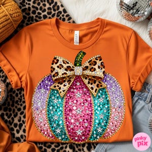Op de afbeelding: Een oranje t-shirt met korte mouwen en een grote afbeelding van een pompoen versierd met veelkleurige strass-steentjes in roze, paarse en turquoise tinten. De pompoen is bekroond met een strik met luipaardprint en een groene strass-steel.