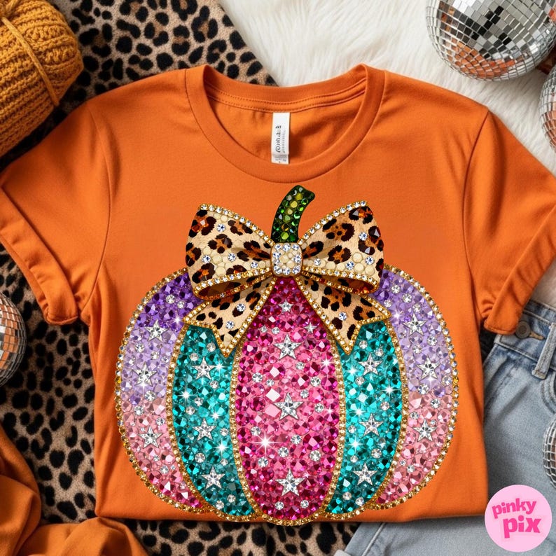 Op de afbeelding: Een oranje t-shirt met korte mouwen en een grote afbeelding van een pompoen versierd met veelkleurige strass-steentjes in roze, paarse en turquoise tinten. De pompoen is bekroond met een strik met luipaardprint en een groene strass-steel.