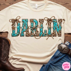 Pu&ograve; includere: T-shirt color crema con la scritta "DARLIN'" in turchese, stampa leopardata e lettere marroni. Le lettere sono decorate con fiocchi di corda e dettagli a stella. Uno stivale da cowboy in pelle marrone e jeans sono visibili.